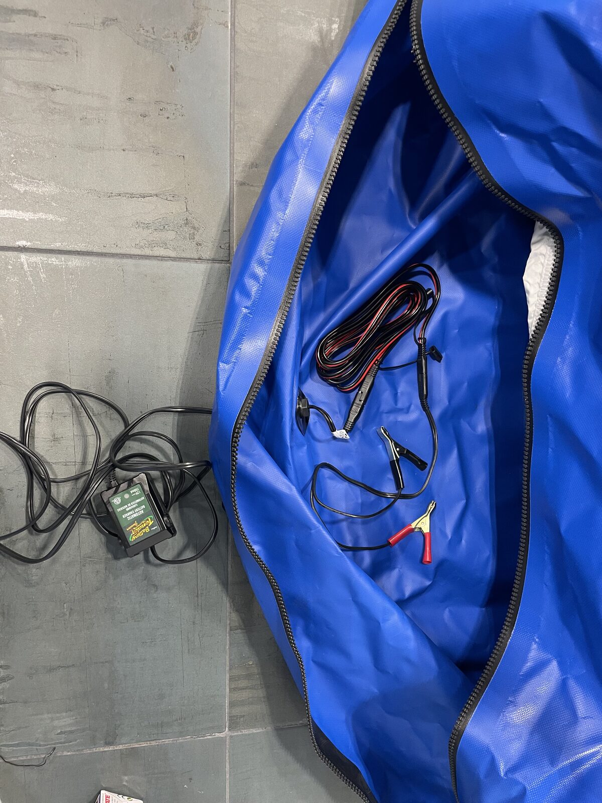 A Battery Tender automatic maintainer sitting on the garage floor outside the sealed blue FloodVault with its cord running to the vault's pass-through port.