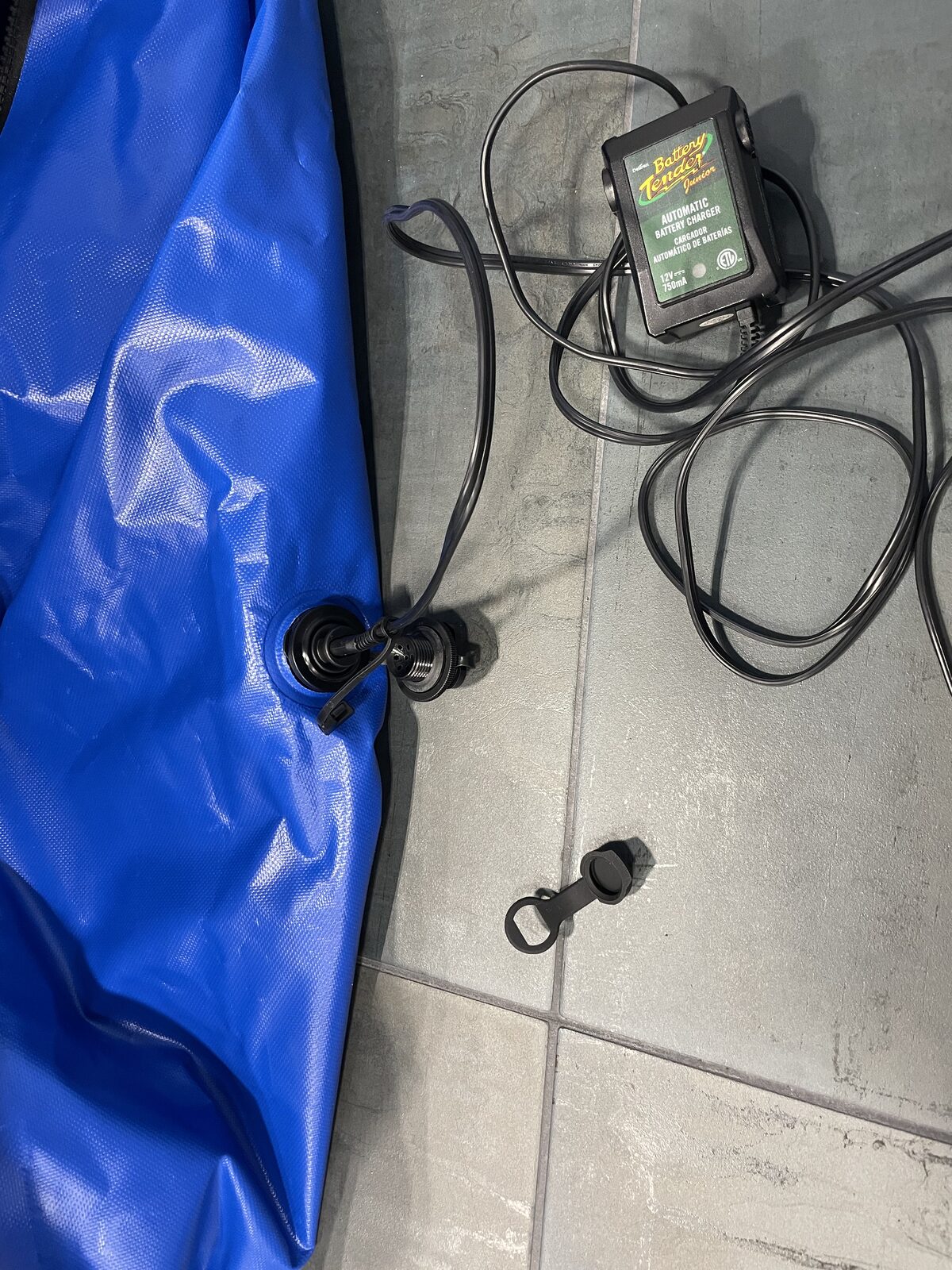 The FloodVault battery charger port — a threaded, gasketed gland welded into the blue PVC envelope with the charger lead passing through, screw cap removed alongside a Battery Tender Junior maintainer.